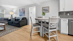 Una sala de estar moderna con un sofá gris, una mesa de comedor con sillas blancas y una cocina con armarios blancos.