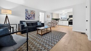 Una sala de estar moderna con un sofá, una mesa de centro y una alfombra estampada. Se ve un comedor con sillas y una mesa al fondo.