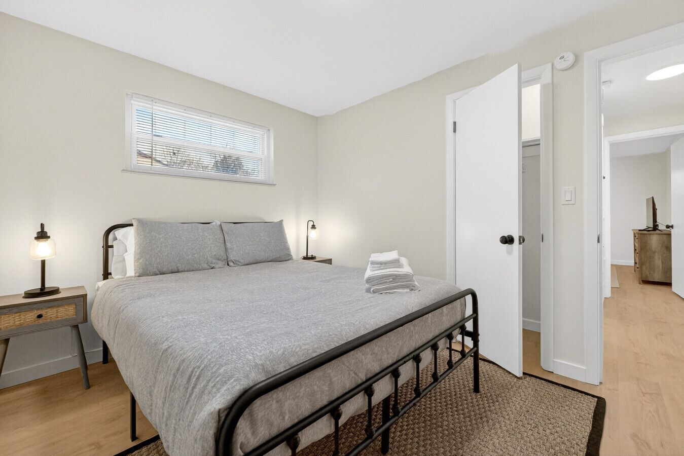 Une chambre à coucher avec un lit, des lampes de chevet, une fenêtre avec des stores, une porte et une petite commode en bois.