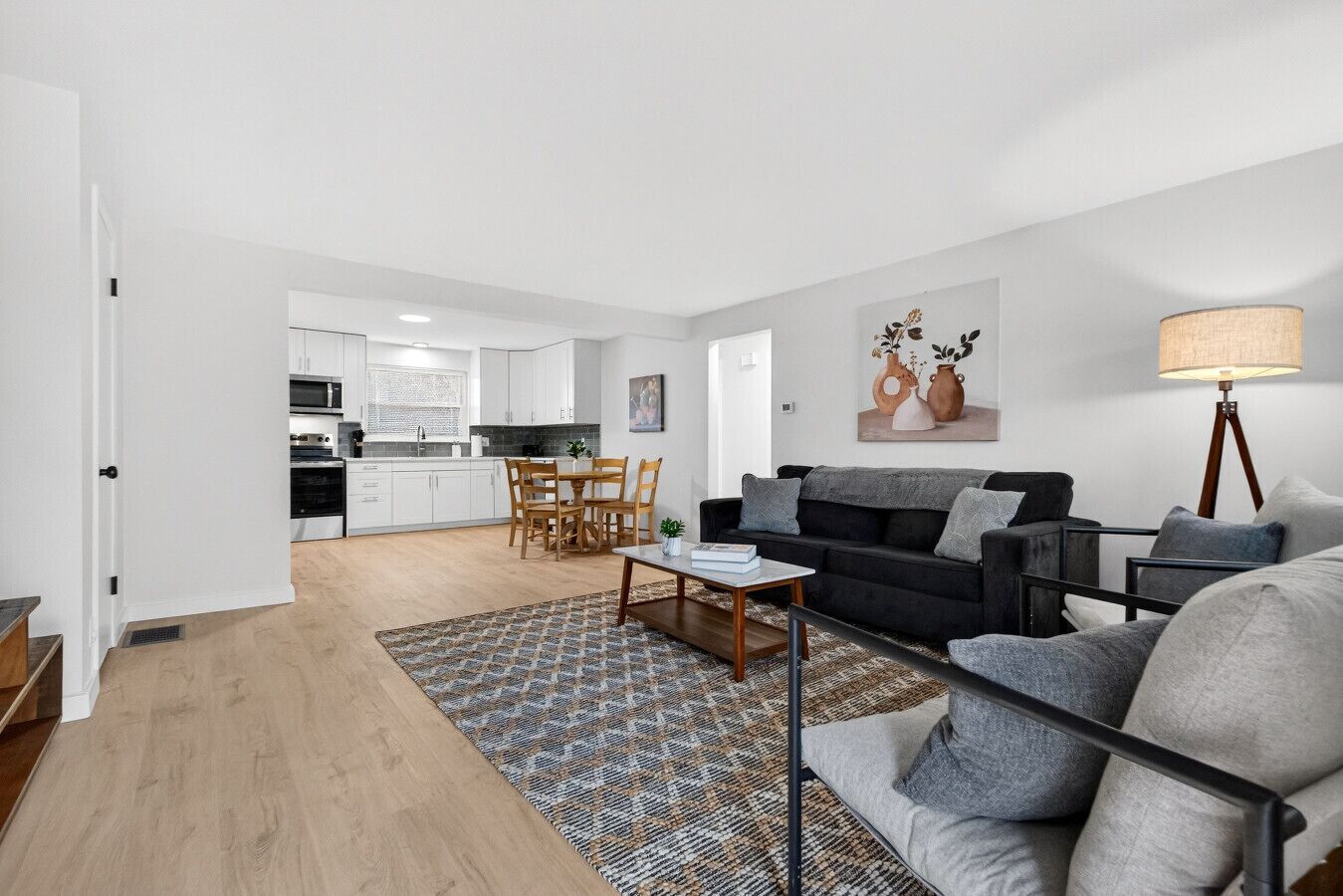 A modern living room with a black sofa, a coffee table, and a patterned rug. There is a dining area with wooden chairs and a kitchen in the background.