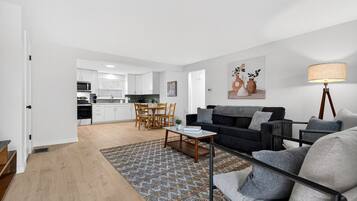 A modern living room with a black sofa, a coffee table, and a patterned rug. There is a dining area with wooden chairs and a kitchen in the background.