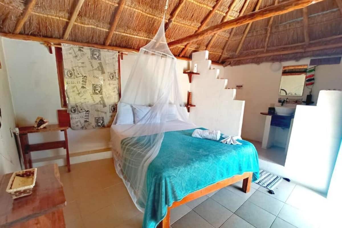 A room with a bed under a mosquito net, a small table, and a bathroom visible through an open door.