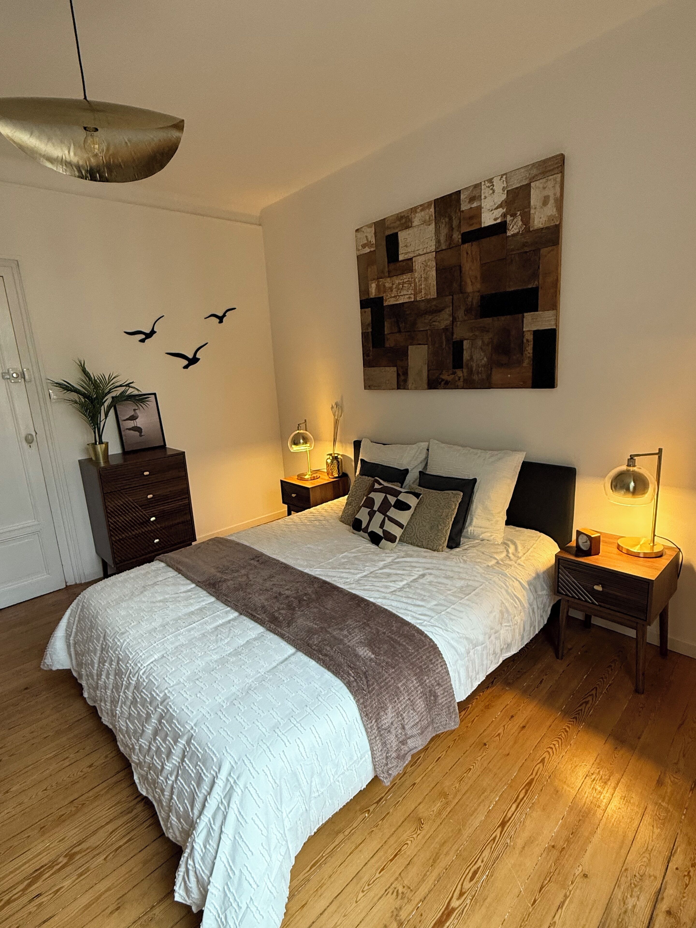 Une chambre avec un grand lit, un plancher en bois, une œuvre d’art abstraite accrochée au mur, un luminaire au plafond, une table de chevet avec une lampe et une petite plante posée sur une commode.