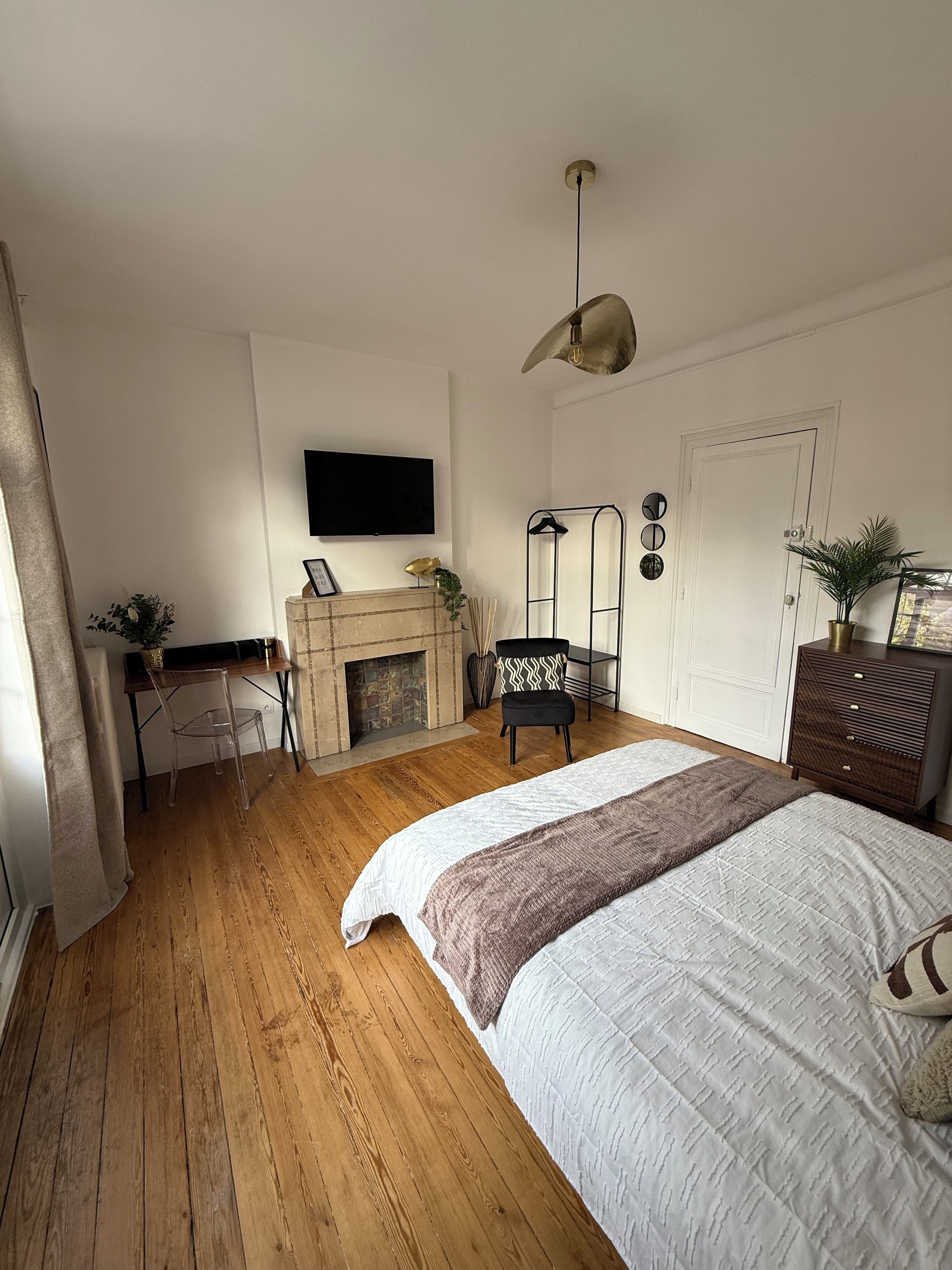 A bedroom with a wooden floor, a bed with a white and brown blanket, a fireplace, a TV mounted on the wall, and a desk with a chair.