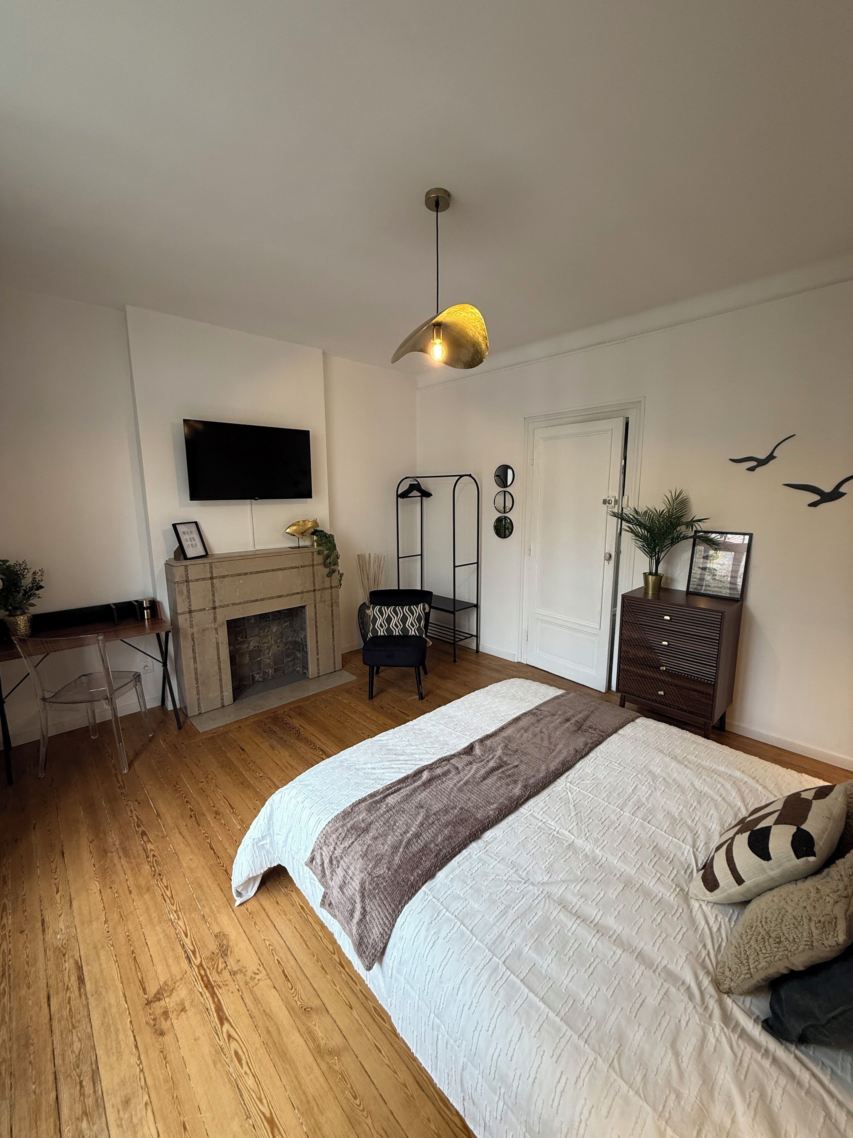 A bedroom with a large bed, a TV mounted on the wall, a fireplace, a desk with a chair, and a dresser.