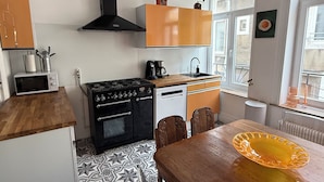 A kitchen with a stove, oven, microwave, and sink. There is a dining table with chairs and a window with a view. - CHAMBRE LE PHARE (Boulogne-sur-Mer)