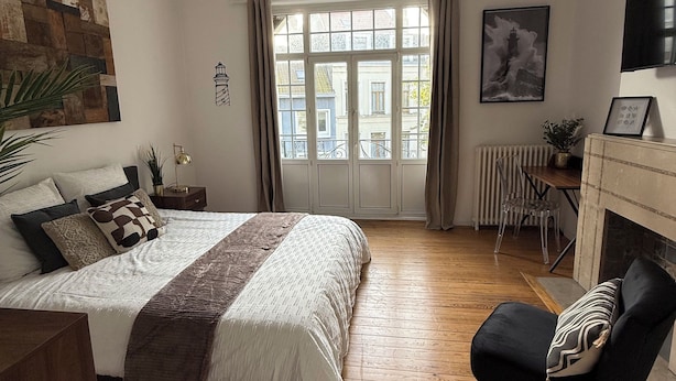A bedroom with a large bed, a chair, a fireplace, and a window with curtains. - CHAMBRE LE PHARE (Boulogne-sur-Mer)