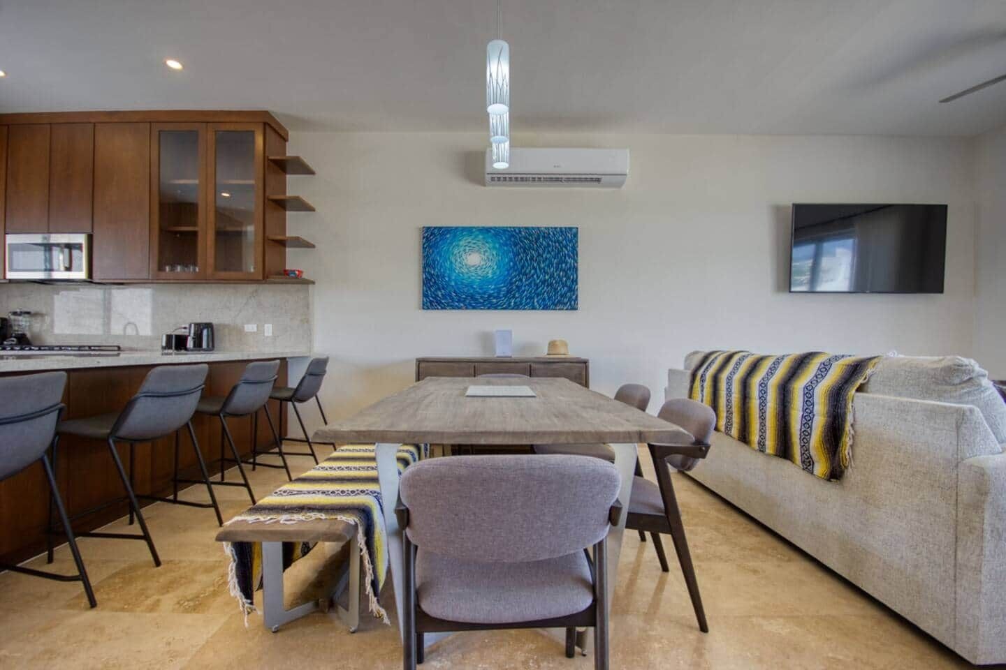 Une cuisine moderne avec des meubles en bois, un coin repas avec une table et des chaises en bois, un canapé recouvert d’un plaid à motifs et un grand tableau abstrait accroché au mur.
