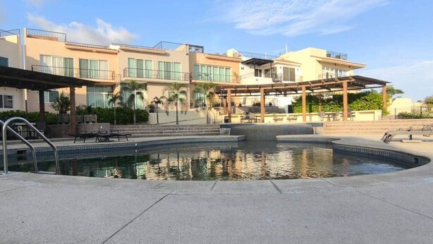 Ein Poolbereich mit überdachter Lounge, ein Gebäude mit mehreren Balkonen und einem wolkenlosen Himmel.
