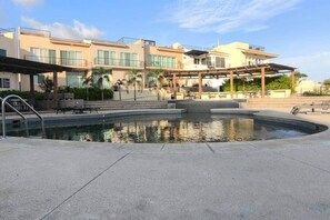 Ein Poolbereich mit überdachter Lounge, ein Gebäude mit mehreren Balkonen und einem wolkenlosen Himmel.