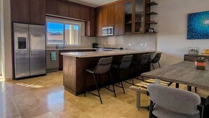 Eine moderne Küche mit dunklen Holzausziehschränken, einem Edelstahlkühlschrank, einer großen Kücheninsel mit Barhocker und einem Esstisch.
