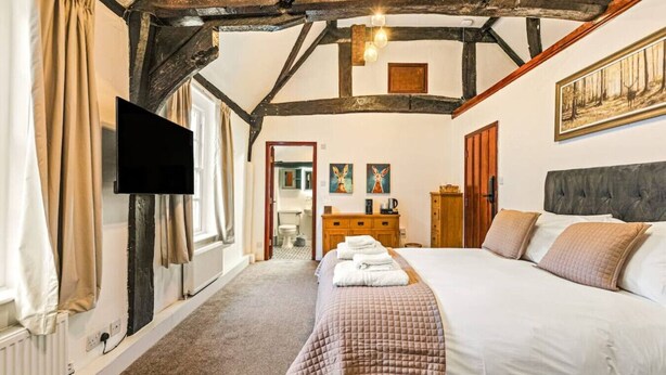 A bedroom with a large bed, wooden beams, a flat-screen TV, and artwork on the wall. - The George Inn Rooms - Room 2 (Great Missenden)