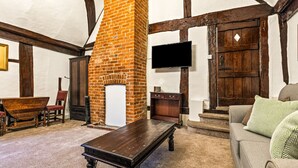 A cozy room with exposed wooden beams, a brick fireplace, a television, and a sofa with patterned cushions. - The George Inn - Room 4 (Great Missenden)
