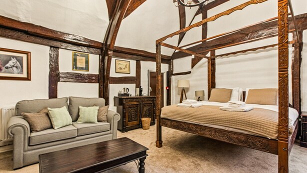 A traditional four-poster bed with a canopy, a sofa, a wooden coffee table, and a dresser in a room with exposed wooden beams and a chandelier. - The George Inn - Room 4 (Great Missenden)
