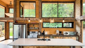 A rustic cabin kitchen with a large island, wooden beams, and large windows offering a view of the forest. - Stunning, Rustic Cottage on Lake of the Woods! (Kenora)