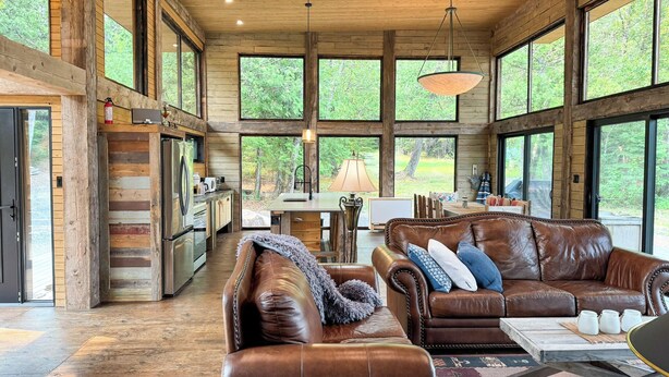 A spacious log cabin interior with large windows, a kitchen, a dining area, and leather sofas. - Stunning, Rustic Cottage on Lake of the Woods! (Kenora)