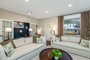 A living room with two sofas, a coffee table with a plant, and a view of a house through a large window. - NEW Comfortable OSU Home • Sleeps 10 • Walk to Campus/Stadium•Free Parking for 4 (Columbus)