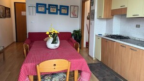 A dining area with a red tablecloth, wooden chairs, and a vase of yellow flowers. There are framed pictures on the wall, a red sofa, and a kitchen area with wooden cabinets and a stove. - Guest Access🌊 Jesolo – Condominio Venezia Apartment✨ Perfect for Families and Groups, Just Steps from the Sea!Welcome to your ideal retreat in Jesolo, where golden sand and crystal-clear sea await you less than 1 minute from your door! 🌅 If you (Lido di Jesolo)