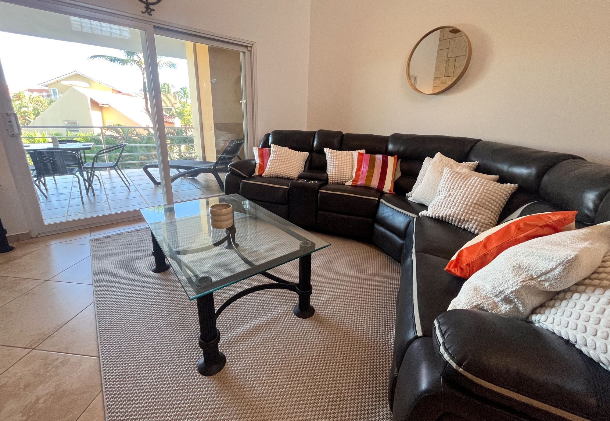 A living room with a glass coffee table, a black leather sofa, and a view of a patio with outdoor furniture.