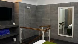 Een moderne badkamer met grijze tegels, een douchecabine met houten frame en glazen wanden, een flatscreen televisie en een grote spiegel.