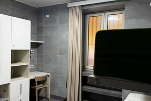 Et lille værelse med grå flisevægge, et vindue med gardiner, en seng og et lille skrivebord med stol.