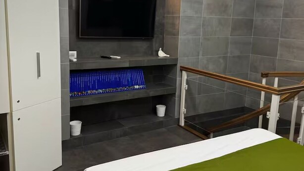 Een moderne hotelkamer met een flatscreen televisie bevestigd aan een grijze muur, een open trap met houten frame en een bed met een groene sprei.