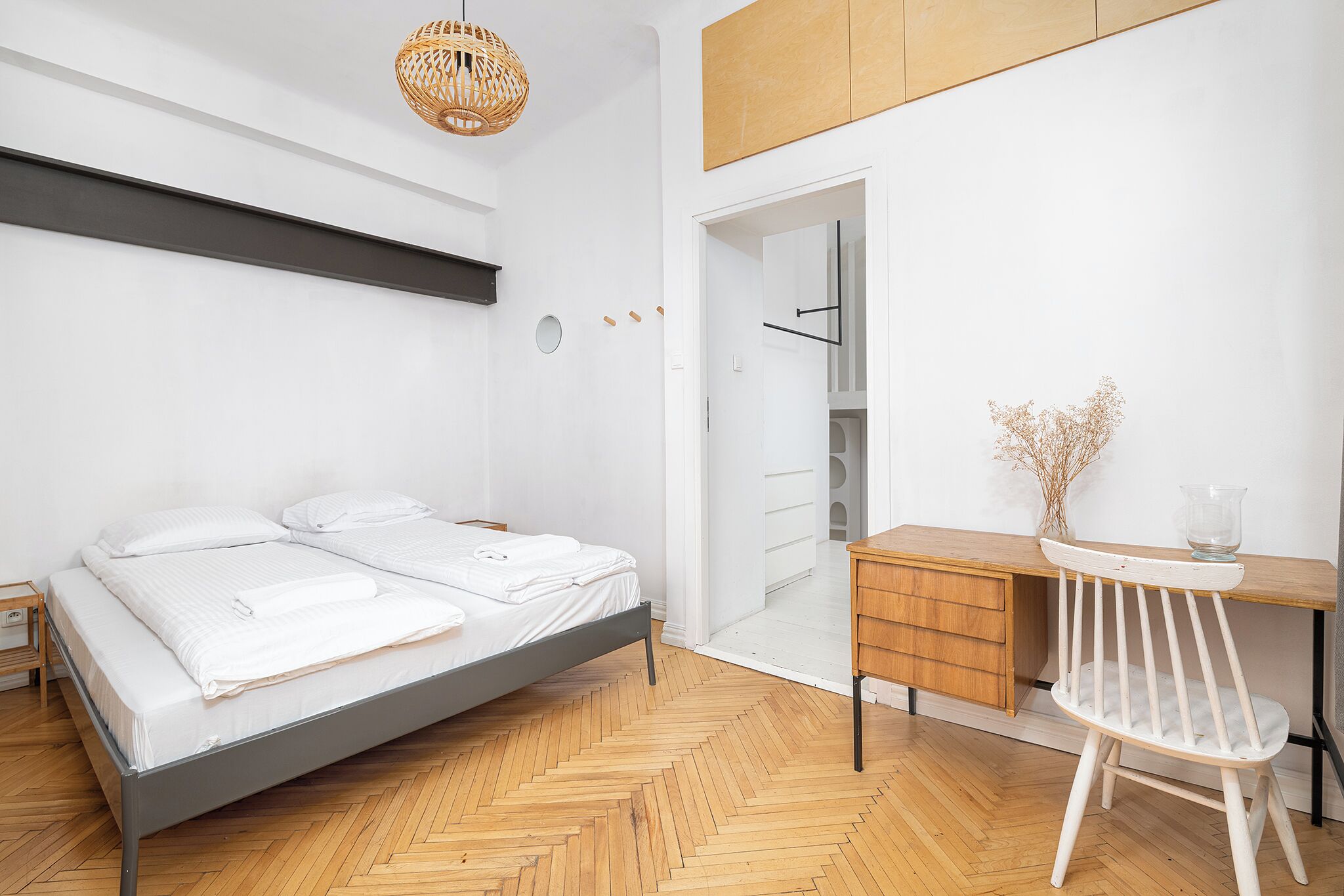 A bedroom with a bed, wooden floor, desk, chair, and a hanging light fixture.