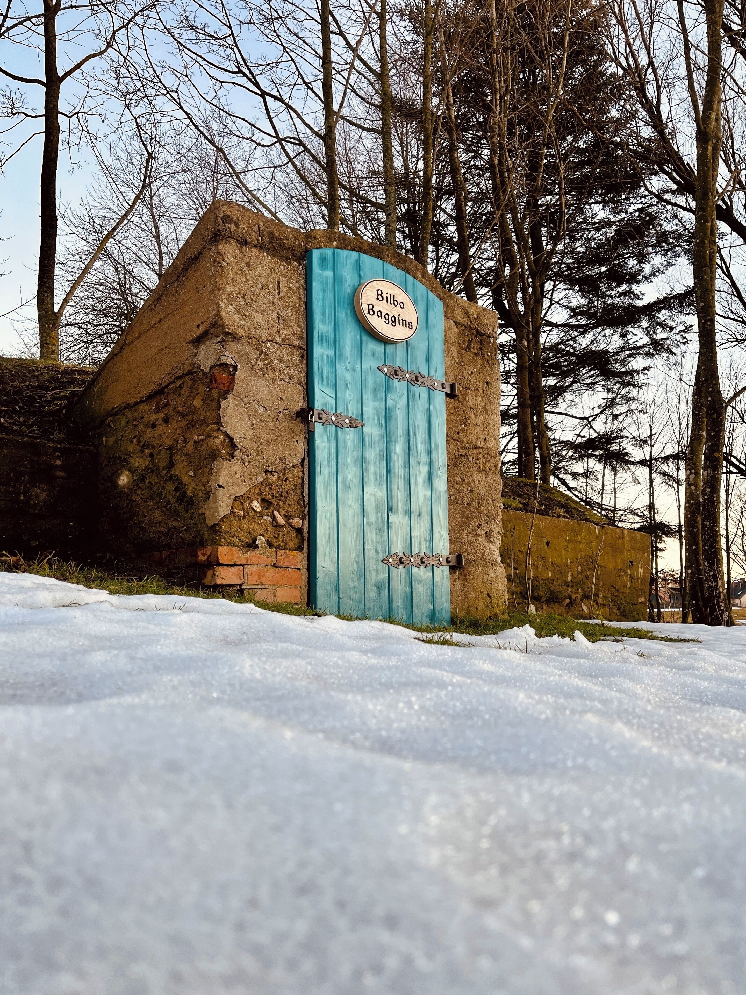 Um edifício rústico de pedra com uma porta turquesa e uma placa com o nome 'Bilbo Baggins'.