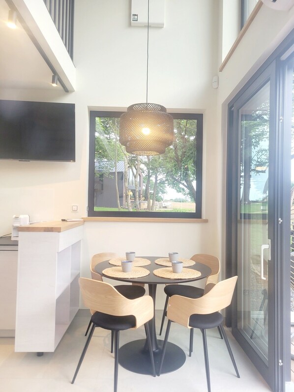 A modern kitchen with a dining area, featuring a large window with a view of trees and a building, a hanging pendant light, and a flat-screen TV mounted on the wall.