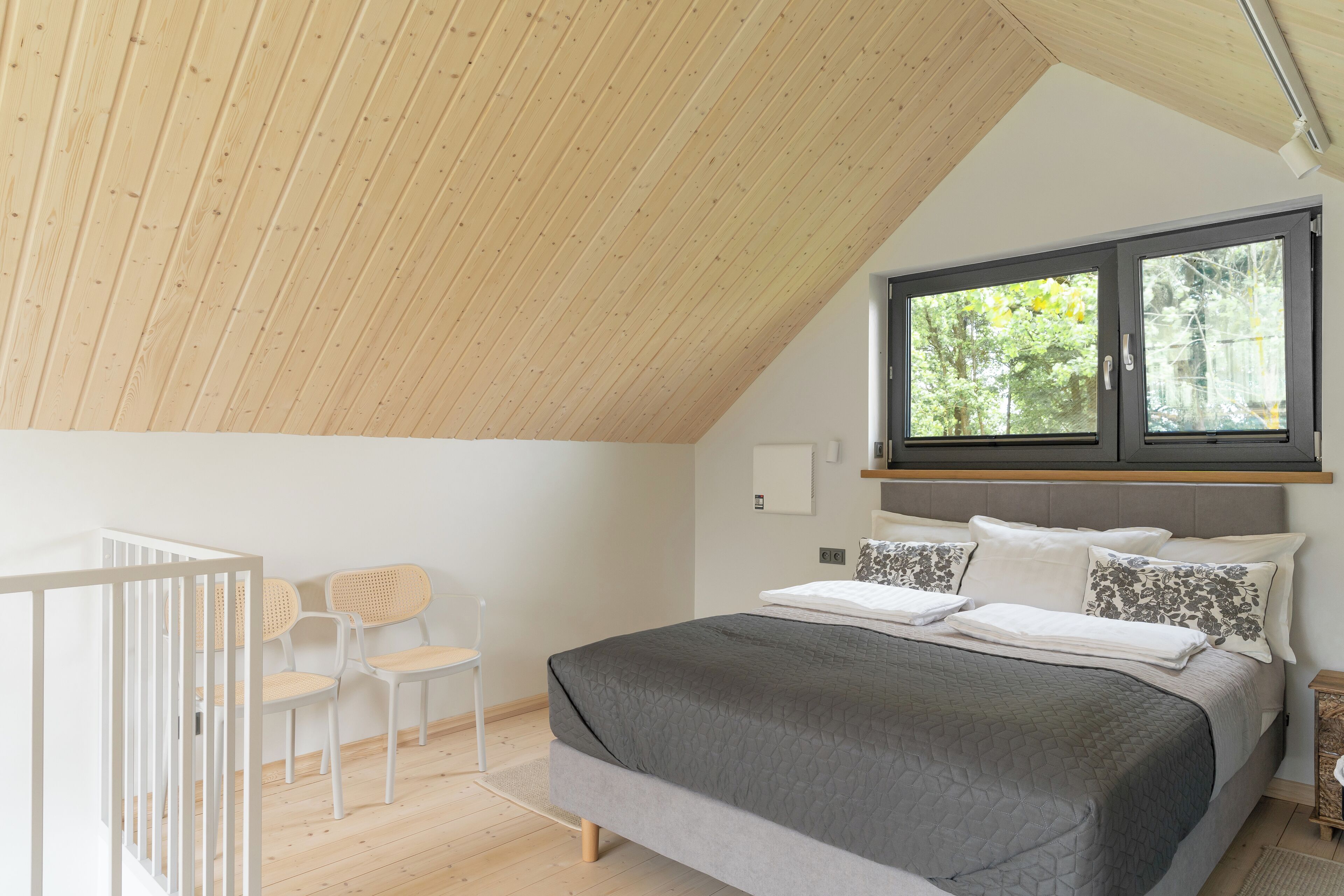 Une chambre avec un plafond en pente, un lit avec une literie grise, deux chaises blanches et une fenêtre donnant sur des arbres.