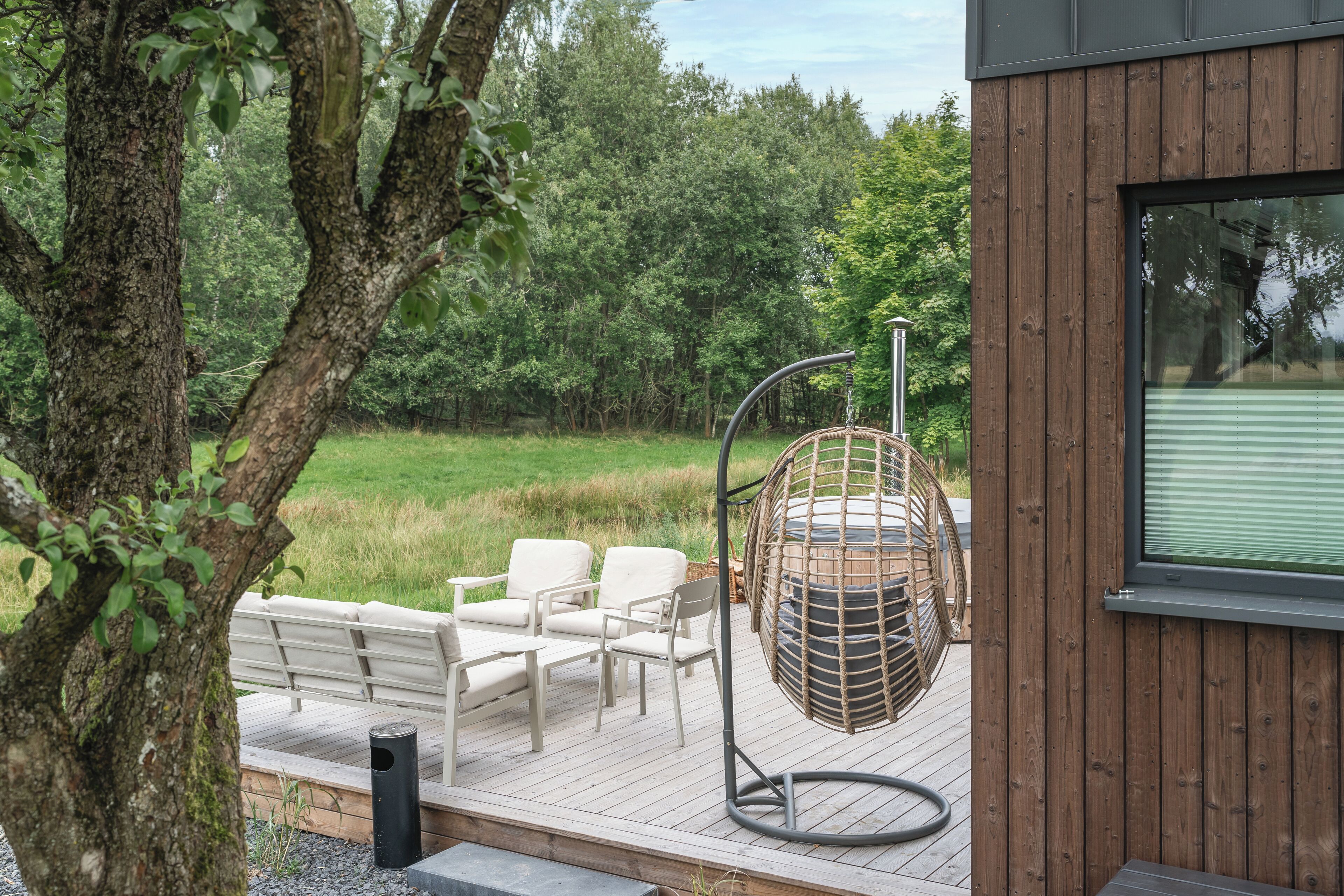 Une terrasse en bois avec des meubles d’extérieur, un fauteuil suspendu et une vue sur une zone boisée.