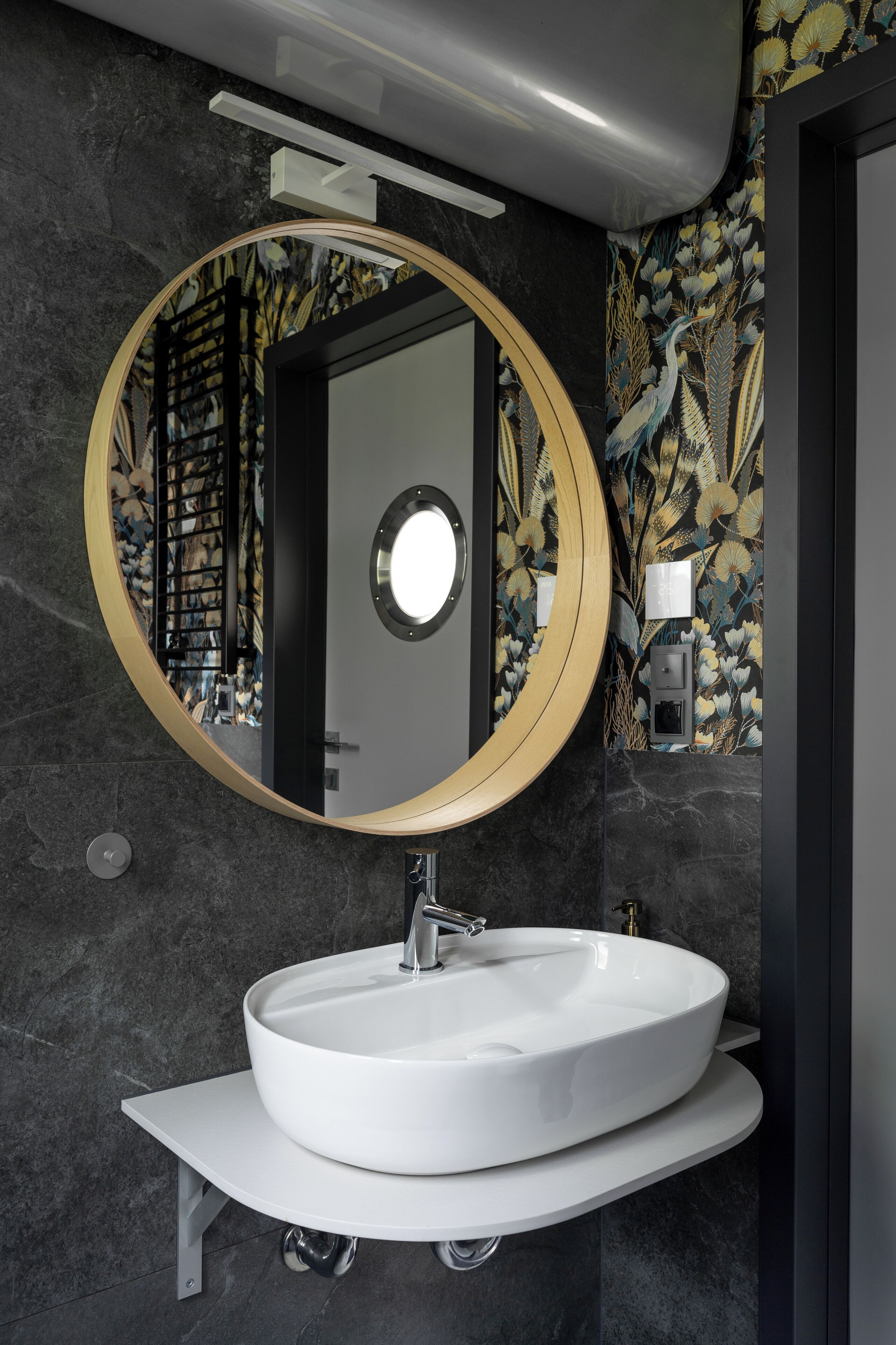 A modern bathroom with a round mirror, white sink, and dark patterned wallpaper.