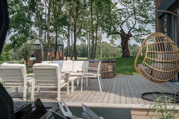 A wooden deck with outdoor furniture, including white chairs and a wicker hanging chair, surrounded by trees. - VILANOVKA (Przodkowo)