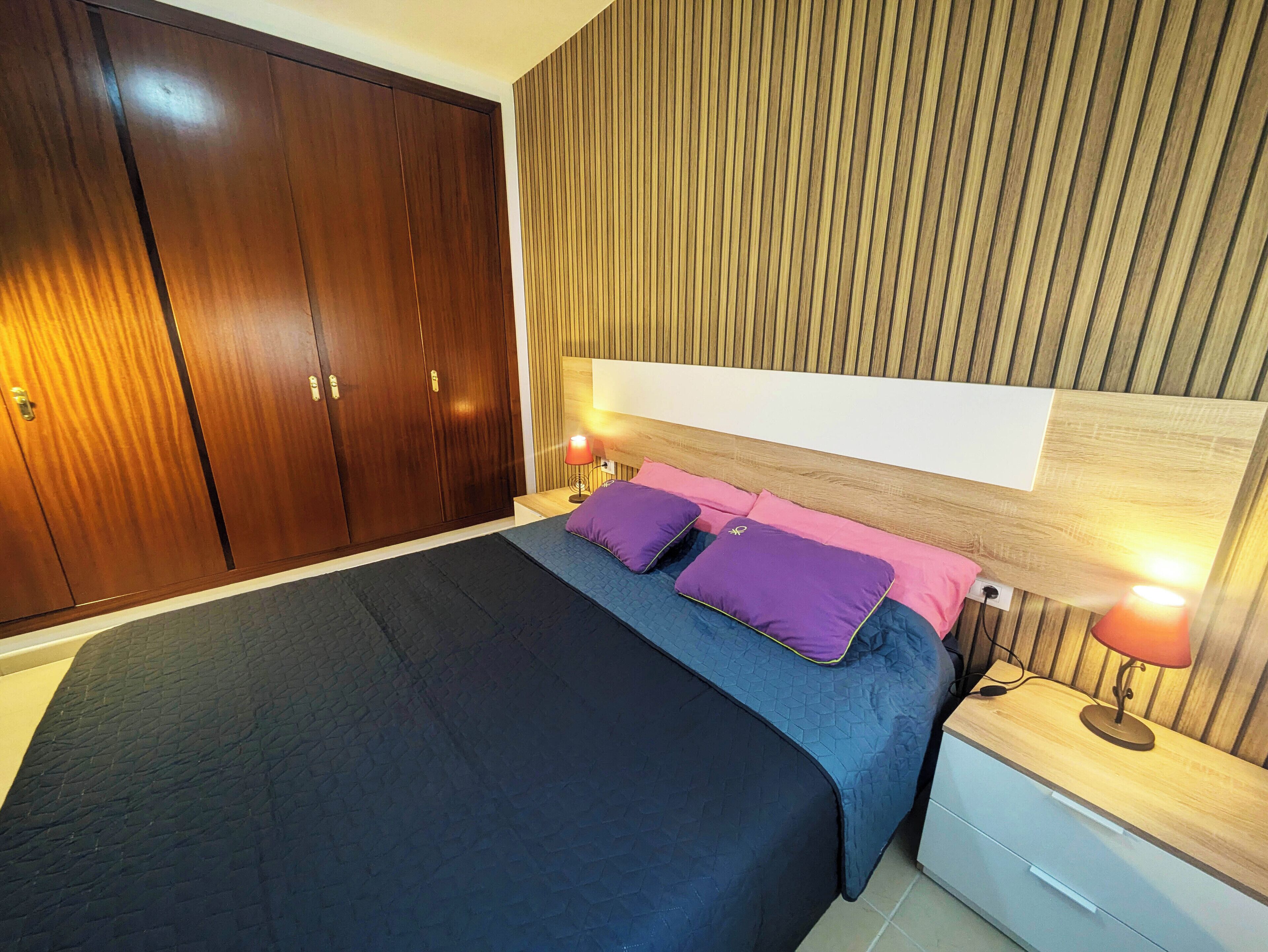 A bedroom with a large bed, wooden wardrobe, and a nightstand with a lamp.