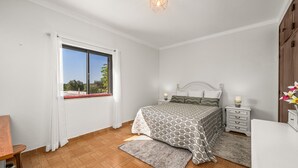 A bedroom with a bed, bedside tables, a window with curtains, a wooden floor, and a ceiling light fixture.