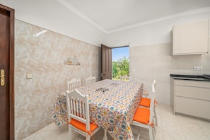 Un coin repas avec une table recouverte d’une nappe à motifs, des chaises blanches avec des coussins orange, un buffet blanc et une vue sur l’extérieur par une porte ouverte.