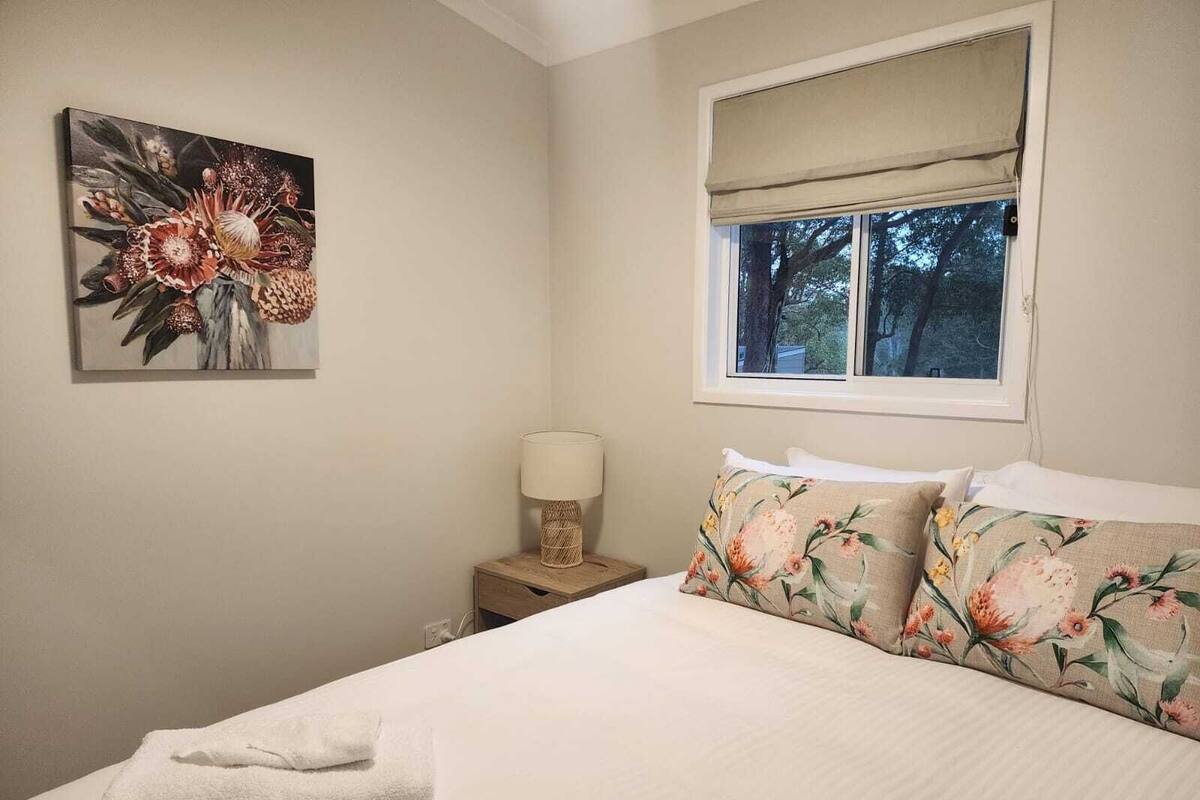 A bedroom with a bed, a nightstand with a lamp, a window with a blind, and a floral painting on the wall.