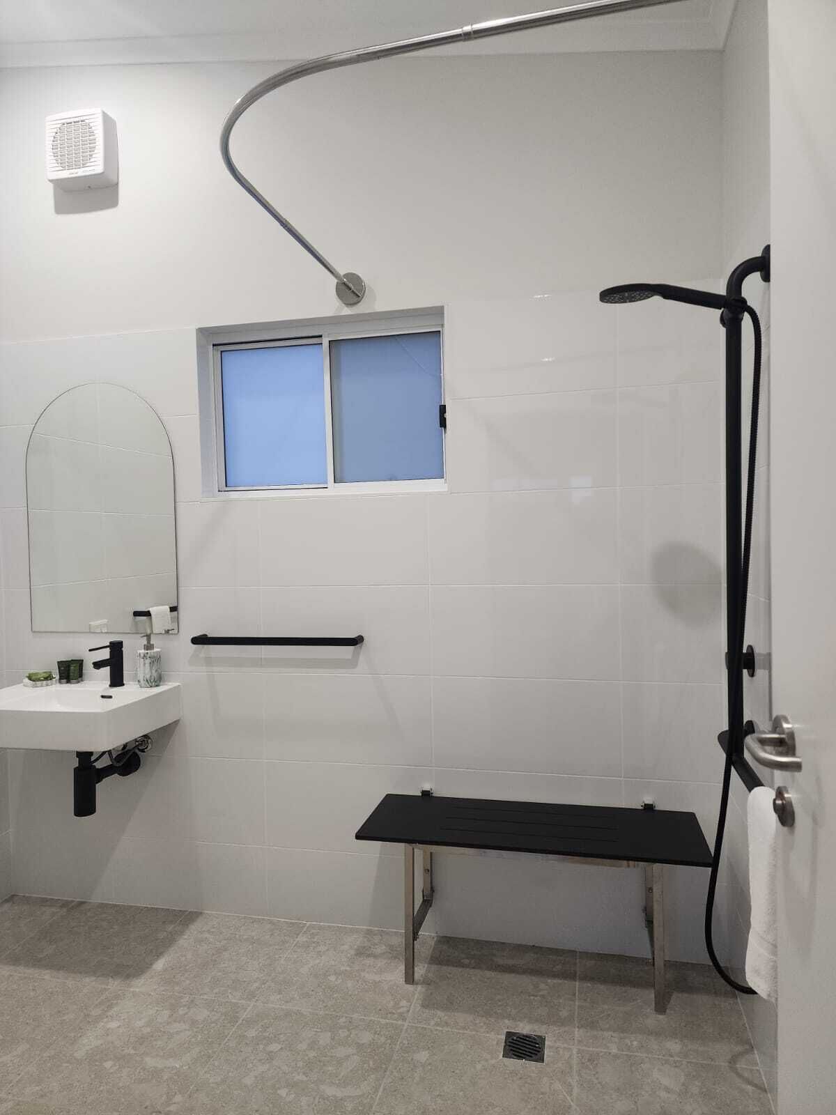 A modern bathroom with a white sink, a mirror, a shower with a handheld showerhead, and a bench.