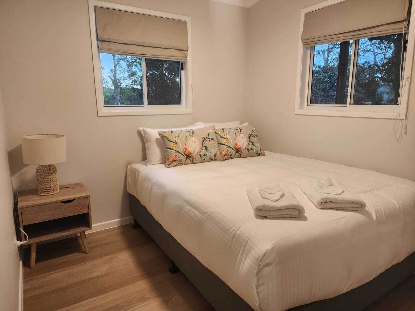 Un dormitorio con una cama, dos ventanas, una mesita de noche con una lámpara y un mueble auxiliar.