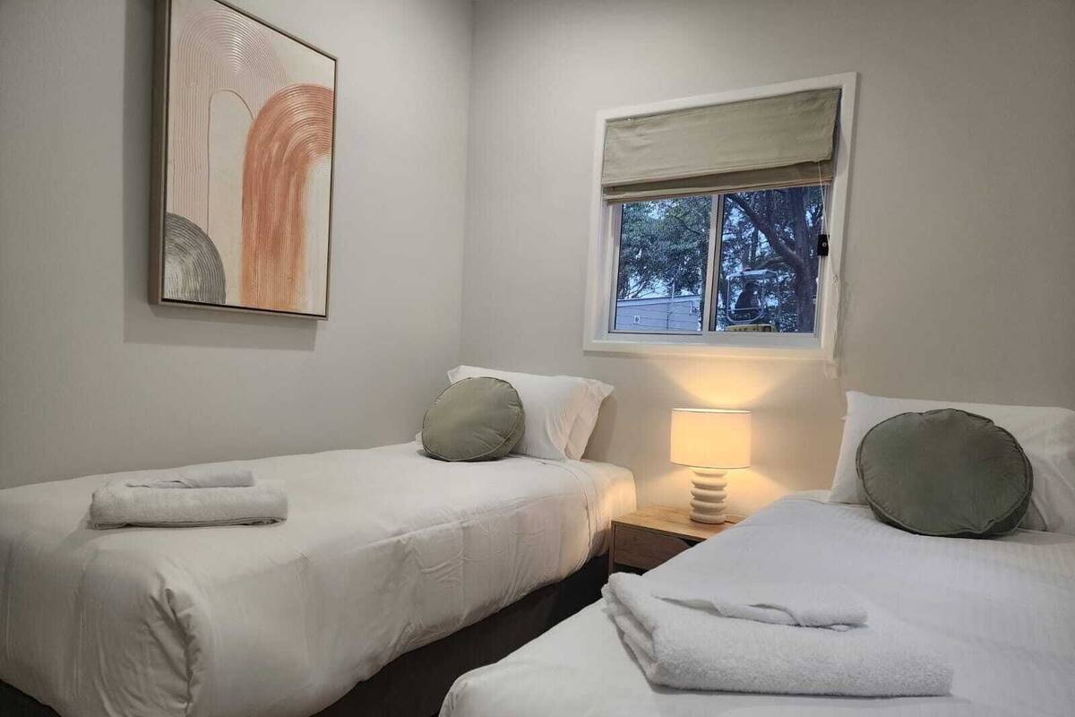A room with two beds, a window with a view, a lamp on a bedside table, and a painting on the wall.