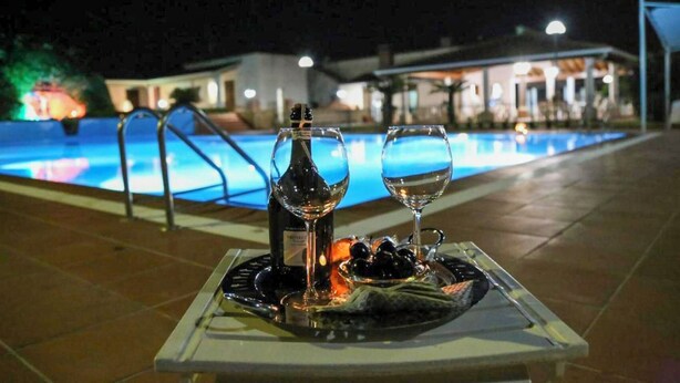 A table with a bottle and glasses on a tray, a pool in the background, and a building with lights. - Villa with private Pool (Ispica)