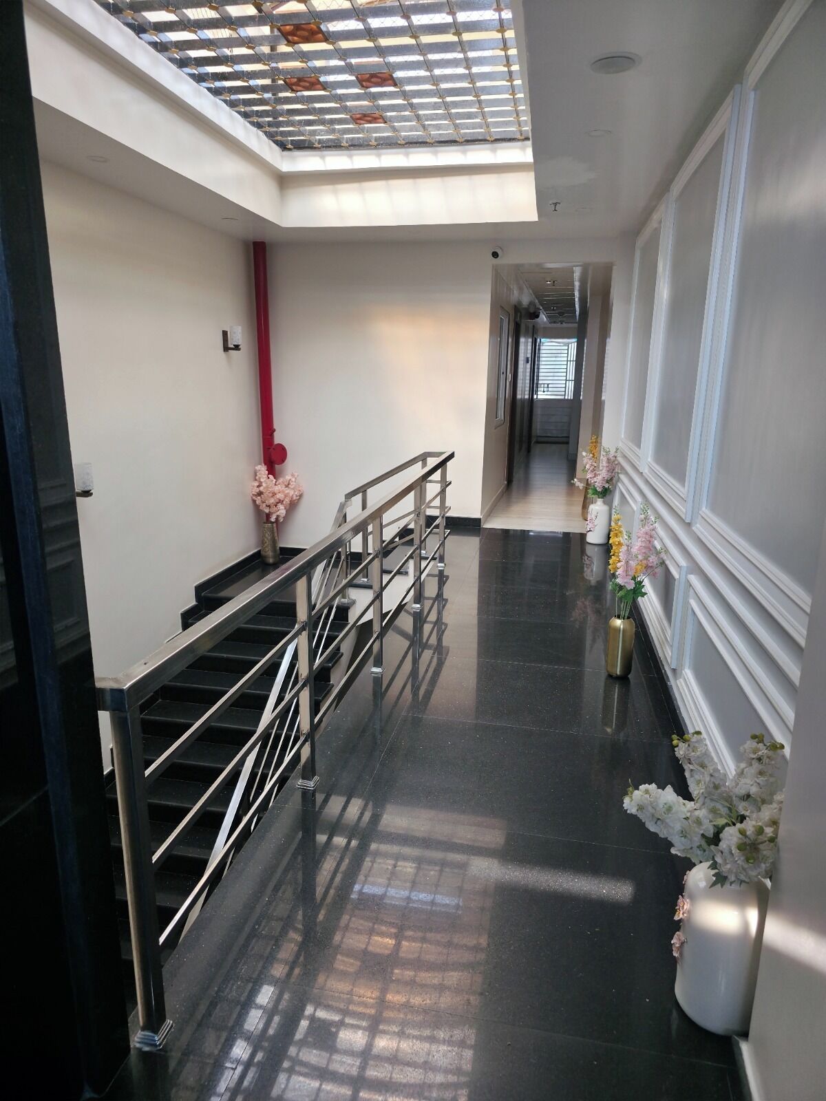 A modern hotel corridor with a glass ceiling, a staircase, and decorative floral arrangements.