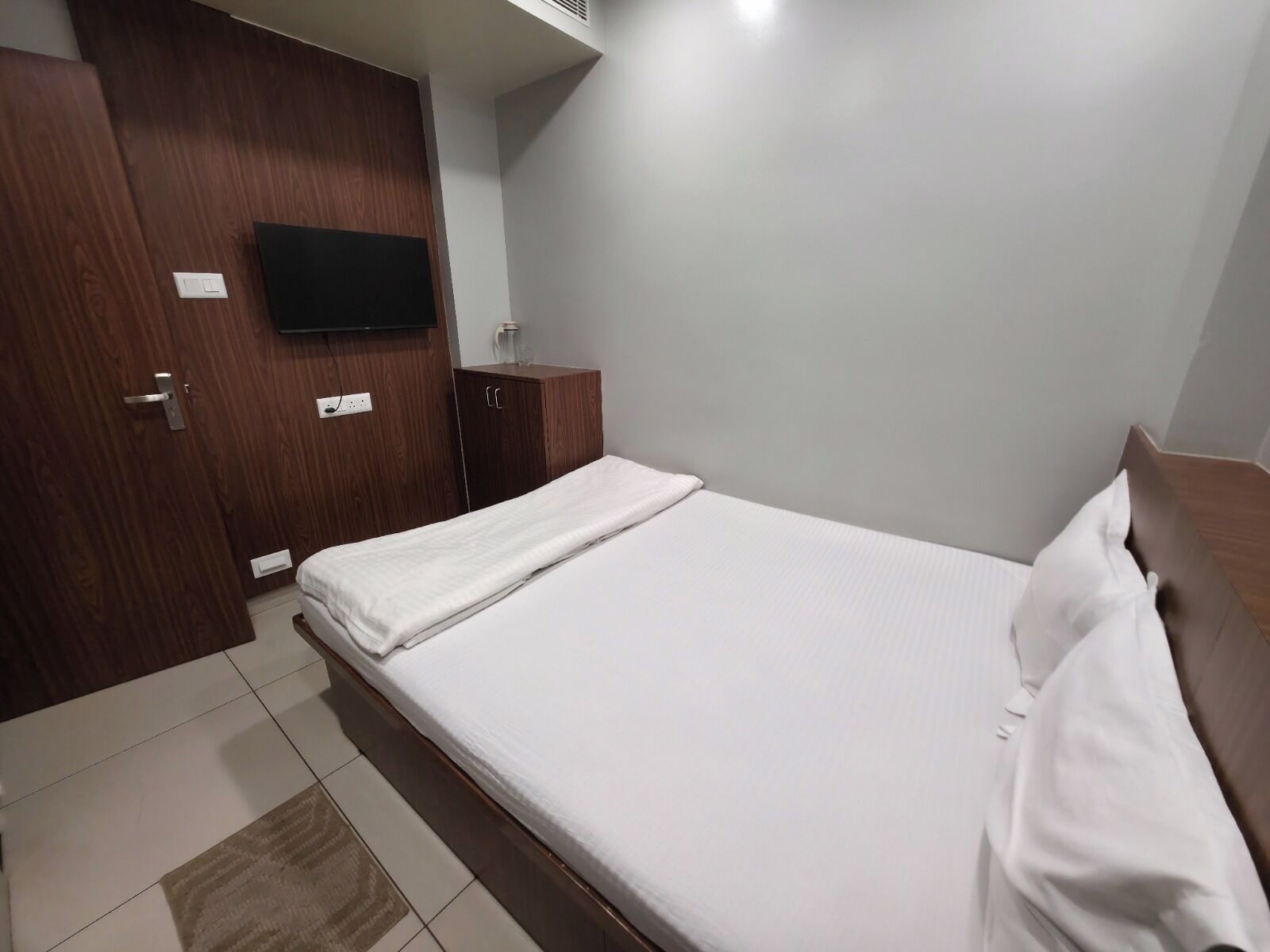 A bedroom with a bed, wooden wardrobe, a flat-screen TV mounted on the wall, and a small bedside table.