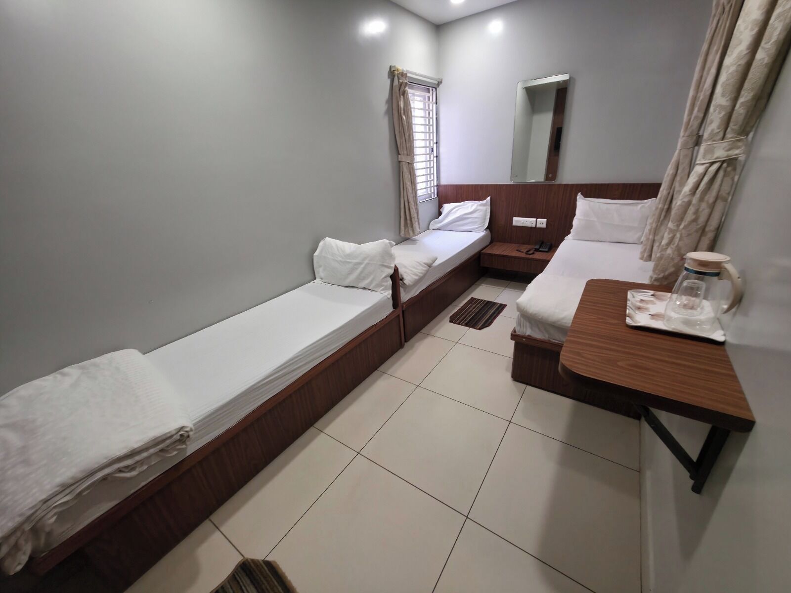 A compact hotel room with two single beds, a small wooden table with a glass container, a bedside table with a phone, and a mirror on the wall.