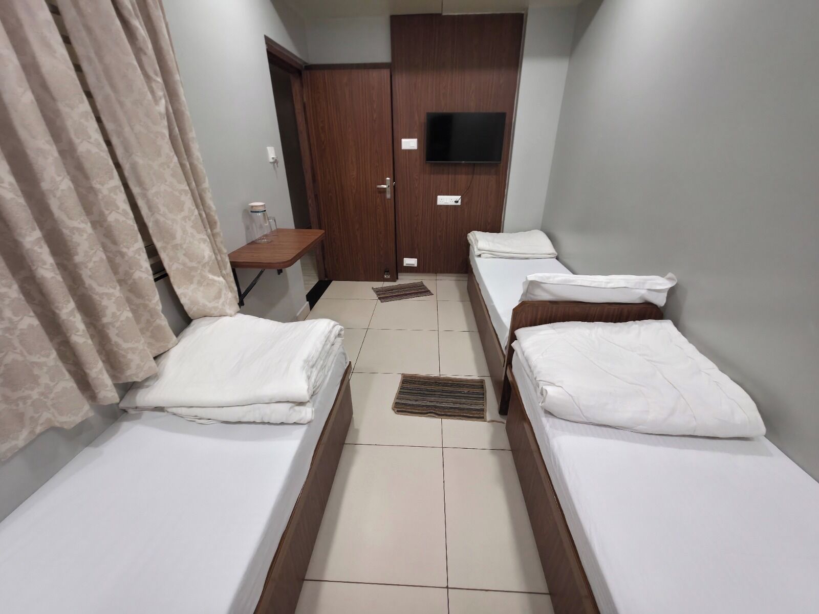 Habitación de hotel con dos camas individuales, un armario de madera, una mesita pequeña y un televisor fijado a la pared.