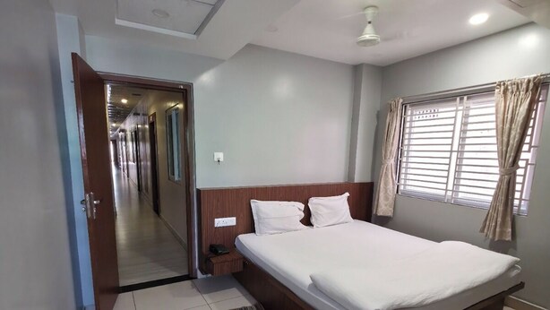 A hotel room with a large bed, a bedside table, a nightstand, a window with curtains, and a door leading to a hallway. - Shree Dham Palace (Puri)