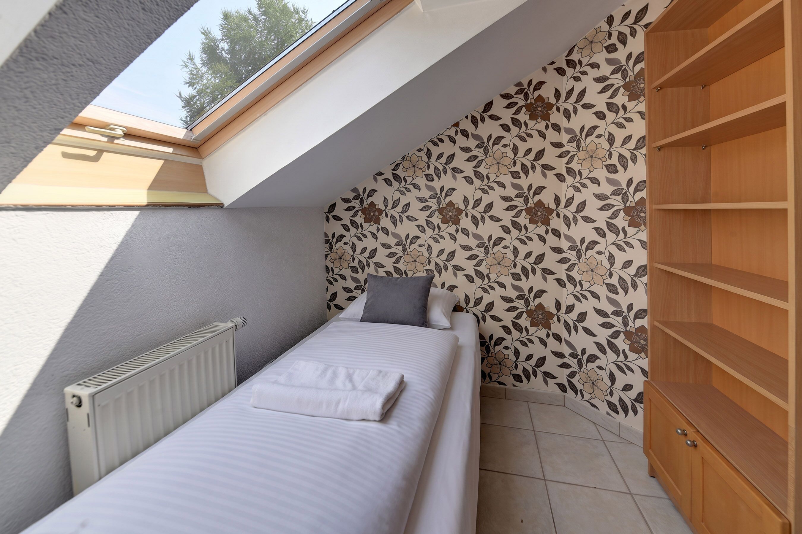 Un dormitorio pequeño con tragaluz, un radiador, una cama con almohadas y un armario de madera.