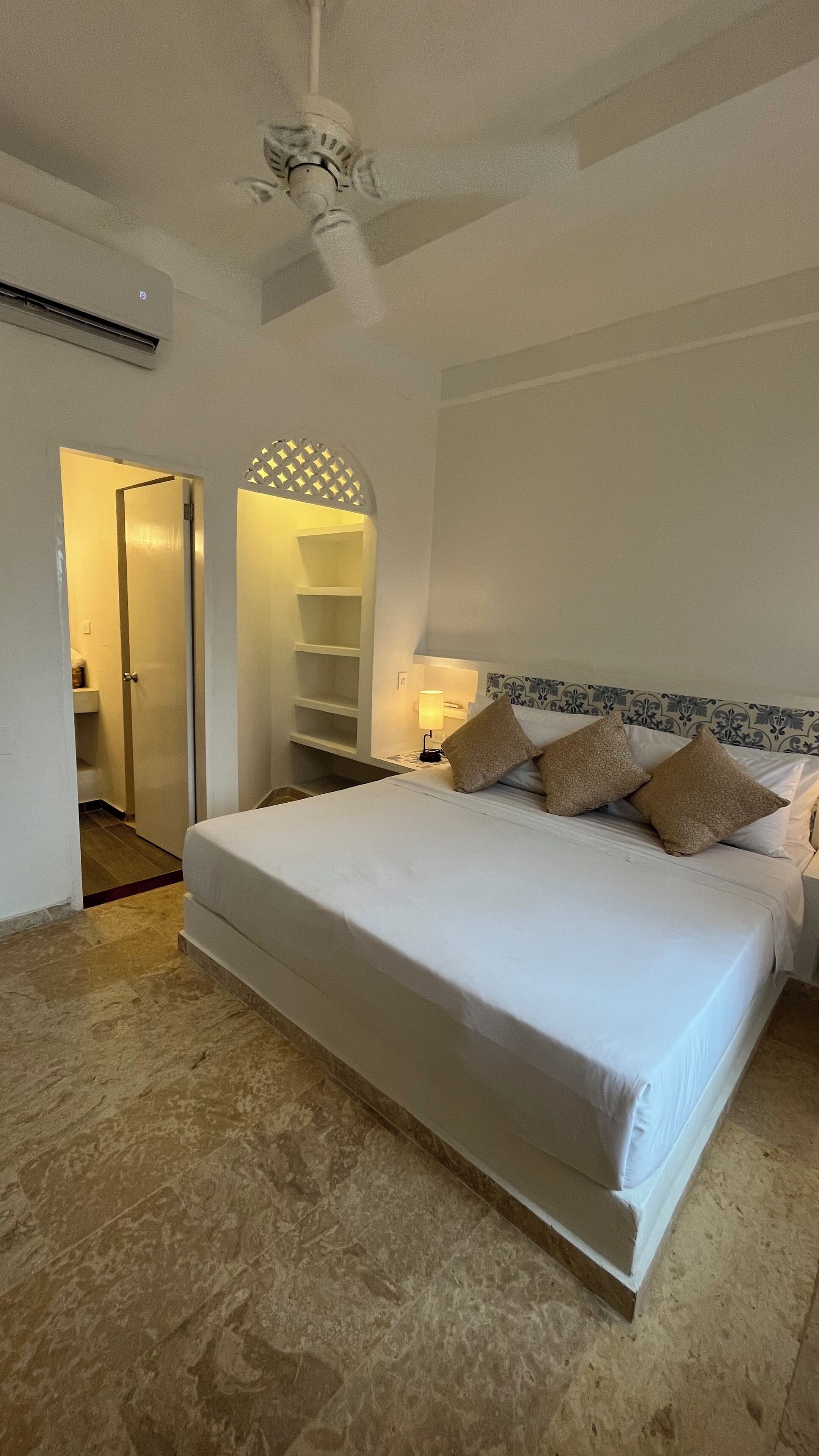 A bedroom with a large bed, a nightstand with a lamp, a ceiling fan, and a door leading to a bathroom.