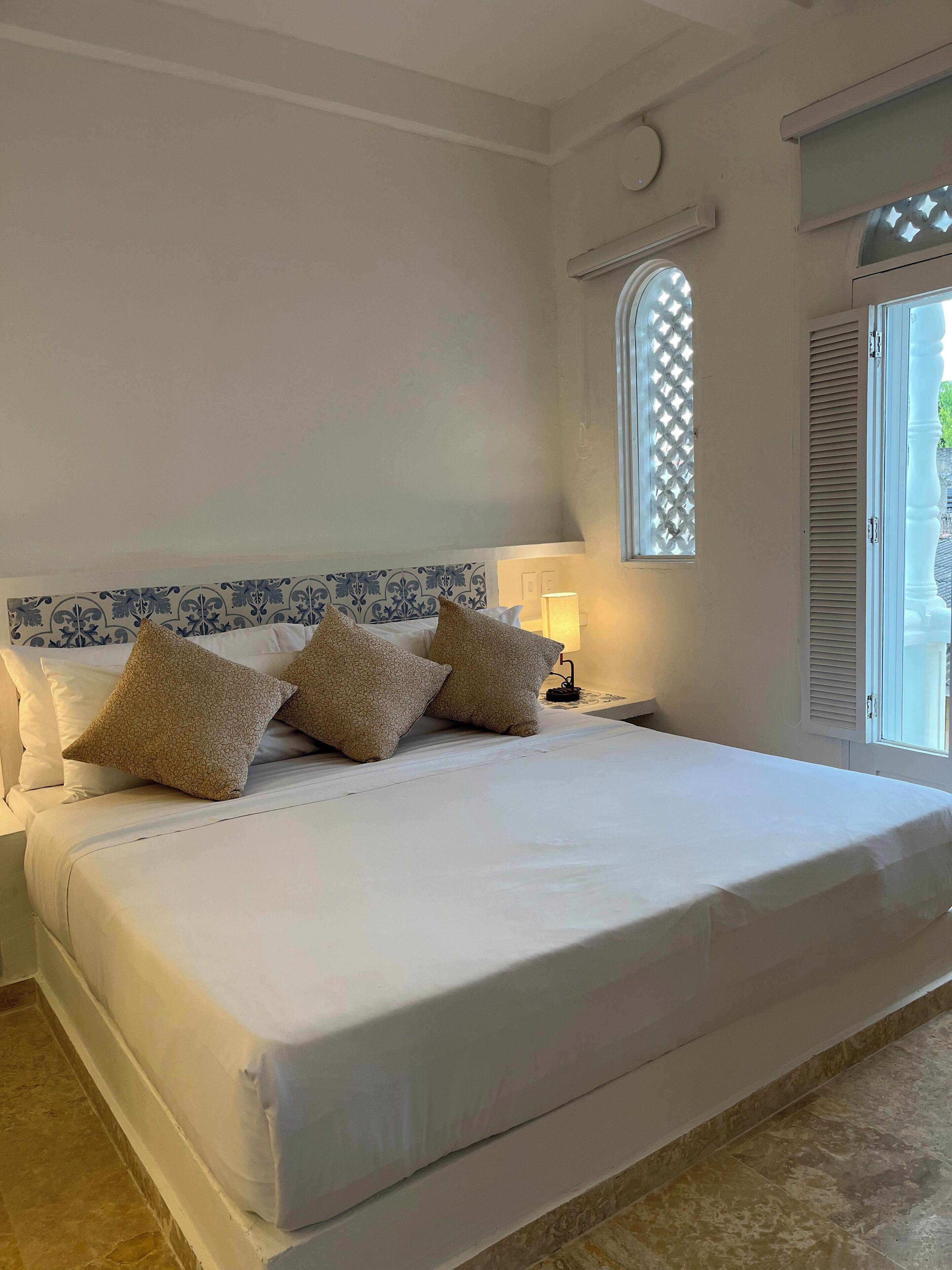 Ein großes Bett mit weißer Bettwäsche und gemusterten Kissen, ein Nachttisch mit Lampe, ein Fenster mit Gittermuster und ein Blick ins Freie.