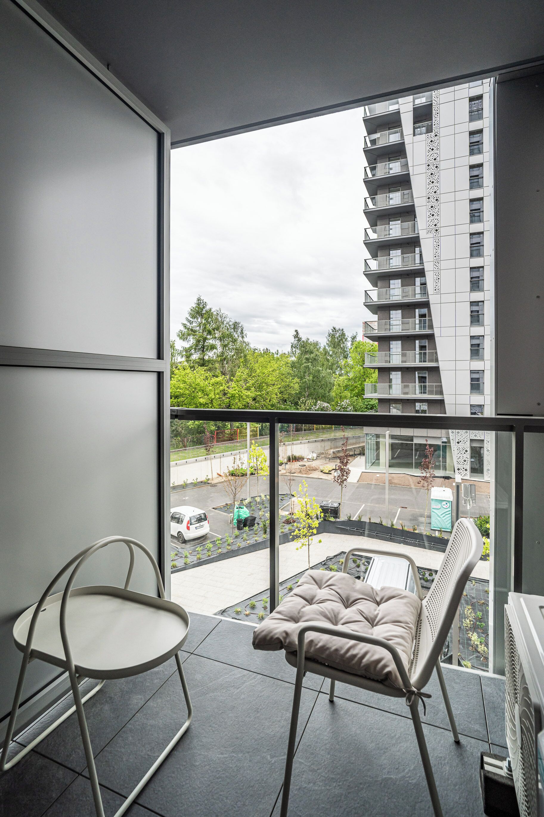 A balcony with two chairs, a view of a parking lot, and a tall building in the background.